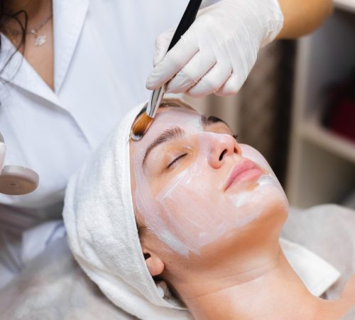 Beautician with a brush applies a white moisturizing mask to the face of a young girl client in a spa beauty salon